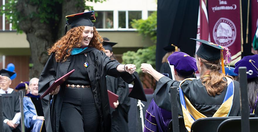Students in cap and gown fist bump after one receives her graduate diploma at SPU